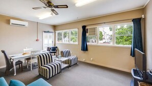 Una habitación con ventilador de techo, una mesa de comedor con sillas, sofás a rayas, un televisor y ventanas grandes con cortinas.