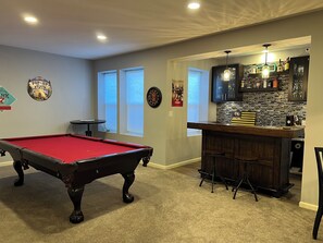 Una mesa de billar en una sala con zona de bar, ventanas y una pared decorativa.