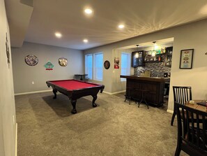 Una mesa de billar en una sala con zona de bar, piso alfombrado y decoración mural.