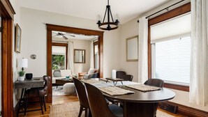 Een eetruimte met een ronde tafel, stoelen en een kroonluchter. Achtergrond is een bank en een raam met jaloezieën.