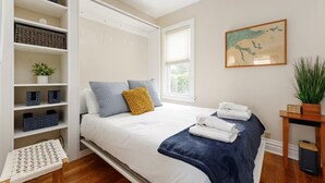 Een slaapkamer met een bed, een inbouwkast, een raam, een schilderij en een potplant.