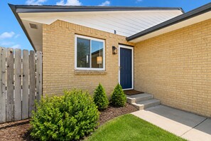 A single-story house with a brick exterior, a blue door, and a small front yard with a wooden fence and shrubs. - Chimney Park Retreat King Suite + Fenced Yard (Windsor)
