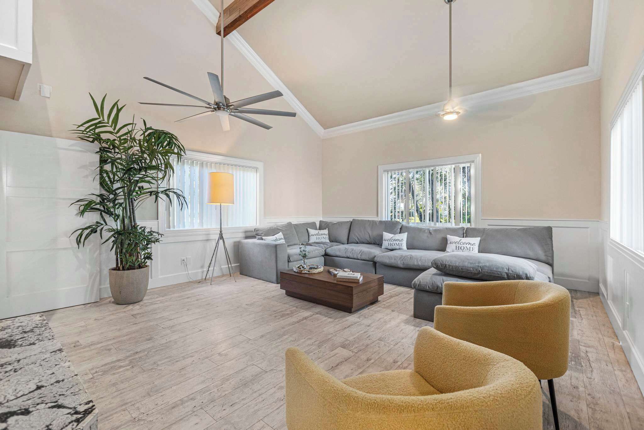 A spacious living room with a large gray sectional sofa, a coffee table, and two mustard-colored armchairs. A potted plant and a floor lamp are also visible.