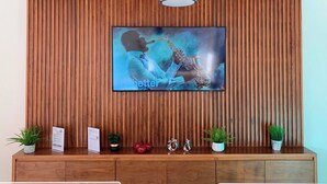 Ein Empfangsbereich mit Wandverkleidung aus Holzlamellen, einem Wandfernseher, der einen Saxophonisten zeigt, und einer Deckenleuchte.