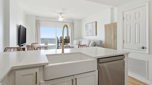 Eine moderne Küche mit weißen Schränken, einer weißen Arbeitsplatte und einem eingebauten Geschirrspüler. Es gibt einen Essbereich mit Korbstühlen und einem Tisch, sowie einen Balkon mit Blick.