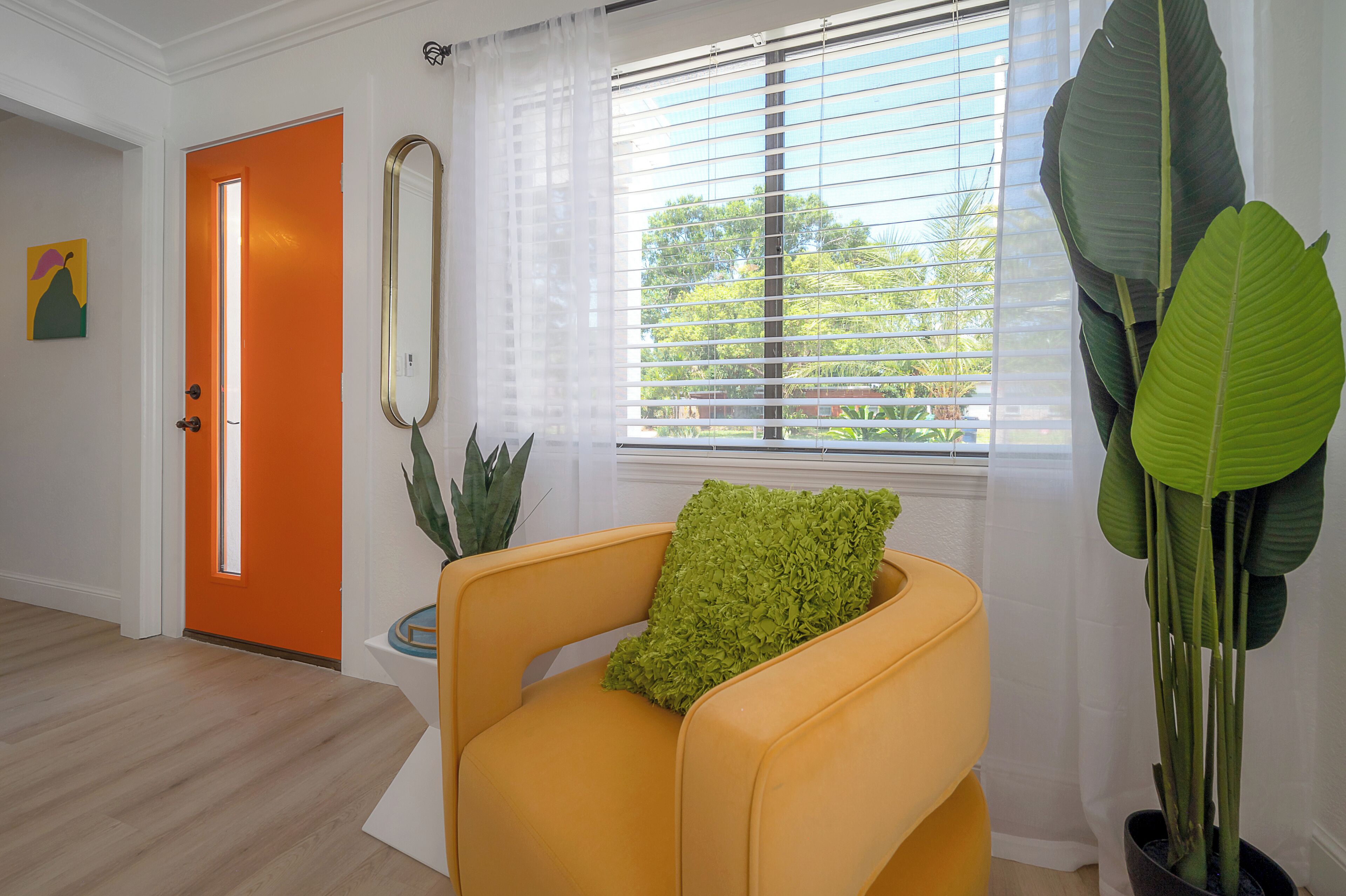 Una habitación con una puerta de color naranja brillante, un sillón amarillo con un cojín verde, una planta grande en maceta y una ventana con persianas.
