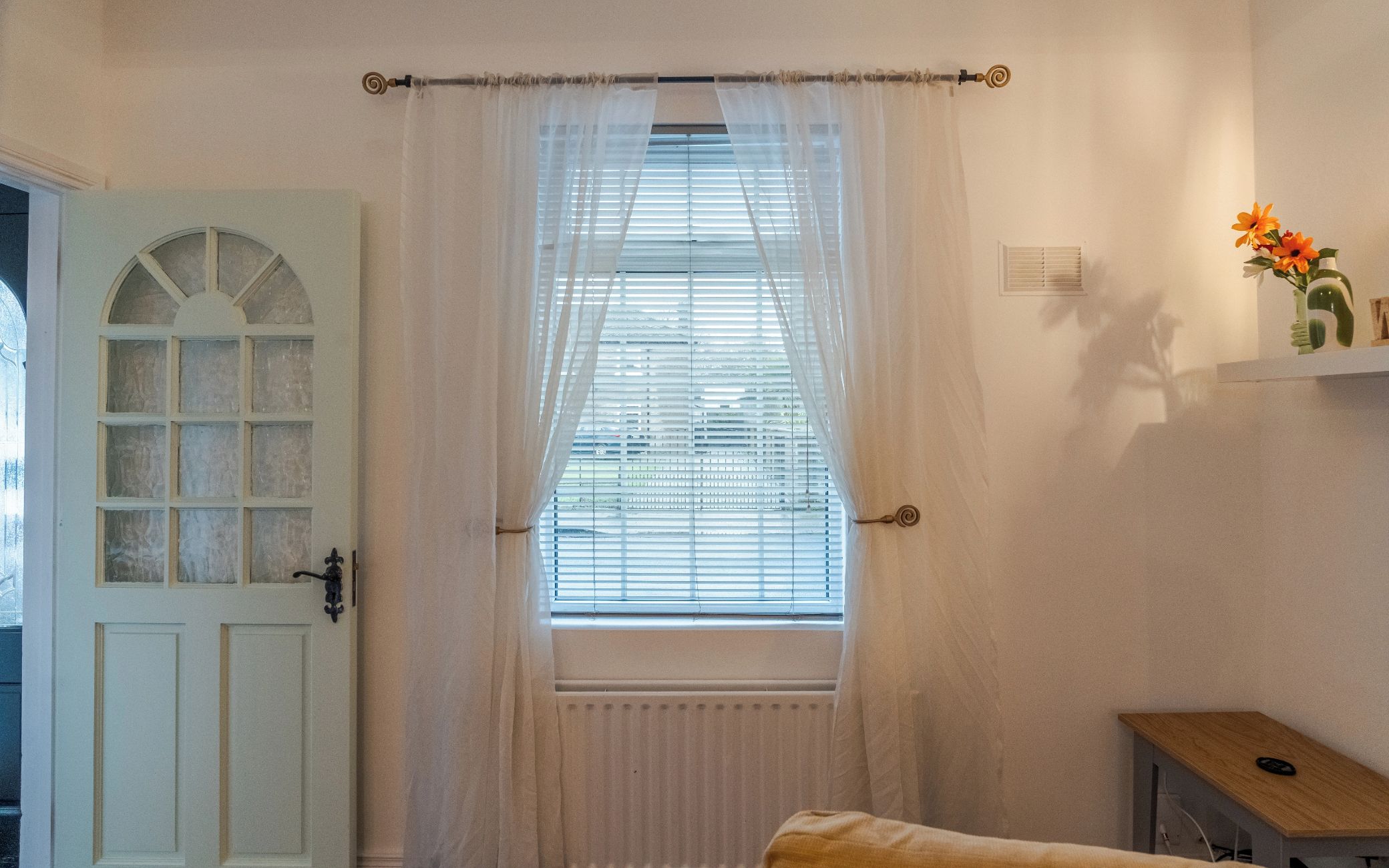 A room with a white door, a window with blinds, a radiator, and a small wooden table with a vase of flowers.