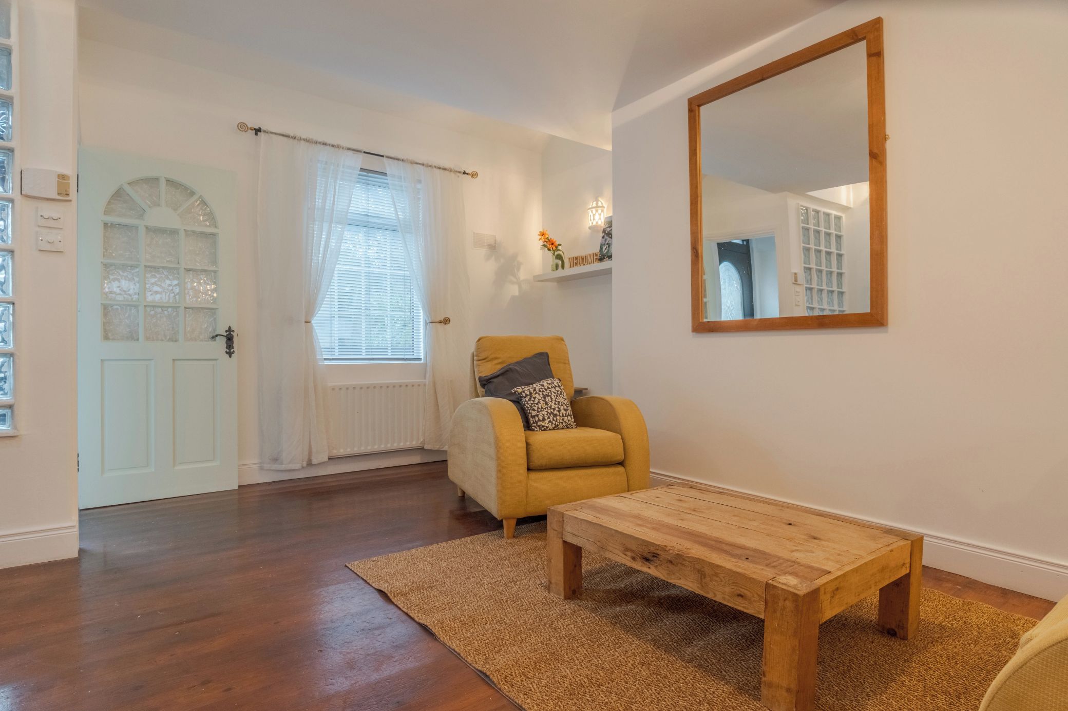 A room with a wooden floor, a yellow armchair, a wooden coffee table, a white door, and a large mirror on the wall.