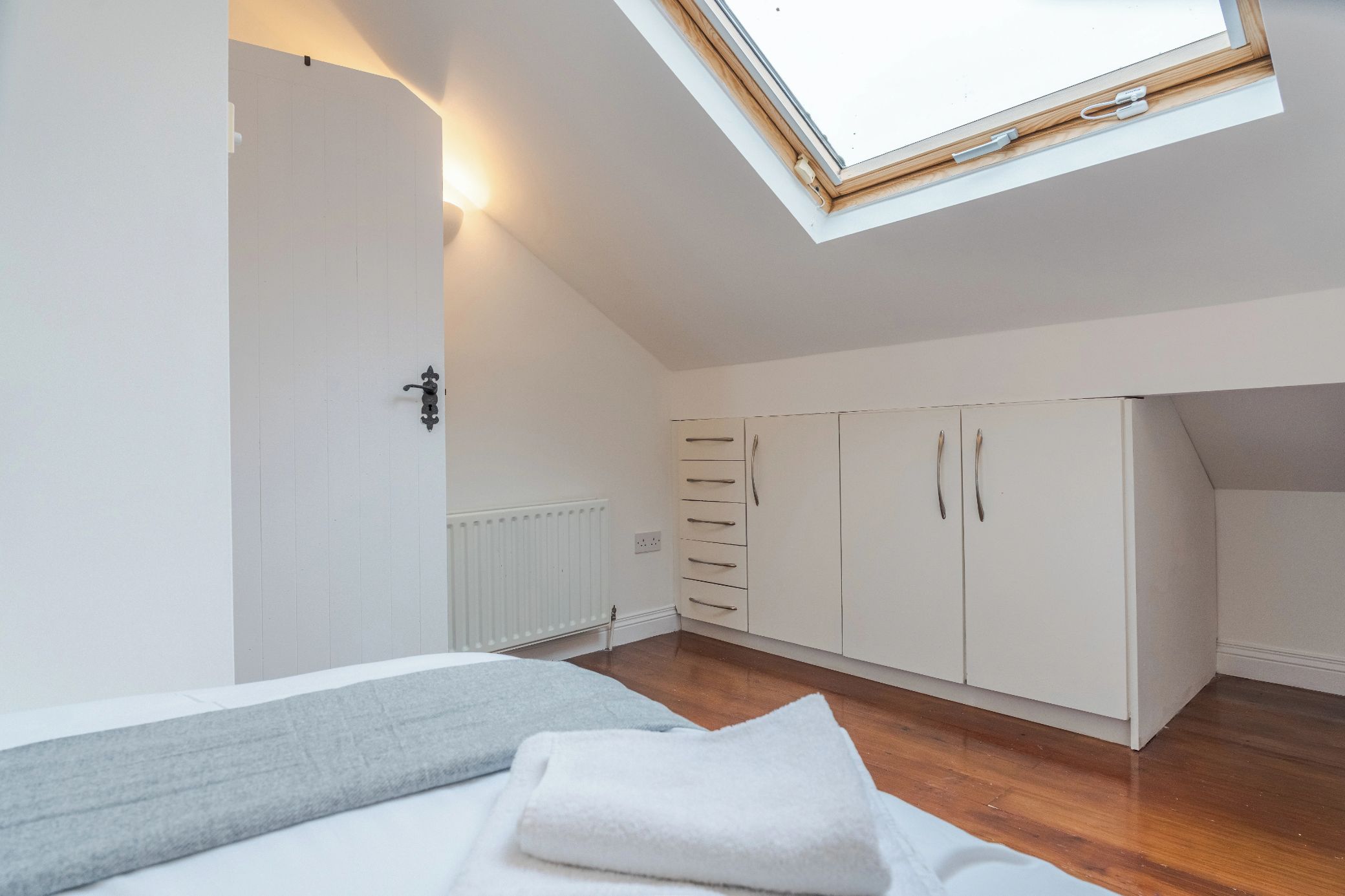 A bedroom with a skylight, a bed with a grey and white blanket, a radiator, and a wardrobe with multiple drawers.