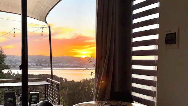 A room with a view of a sunset over a body of water, a table with chairs, and a large window with blinds. - pro estancia 1 (constitución)