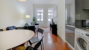 Eine moderne Küche mit integriertem Waschmaschinenanschluss, ein Essbereich mit rundem Tisch und Stühlen sowie ein Wohnbereich mit Sofa und Lampe.