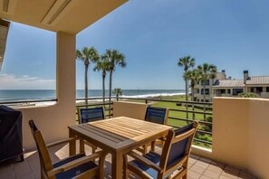 Een balkon met een houten tafel en stoelen, met uitzicht op een strand met palmbomen en een zeezicht.
