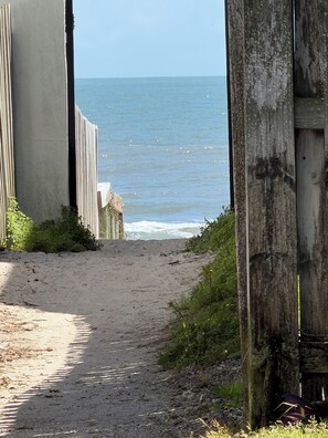 Ein sandiger Pfad führt zum Meer, umgeben von Holzgebäuden und Vegetation.