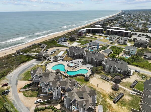 An aerial view of a coastal residential area with a large swimming pool, beachfront properties, and a mix of single and multi-story houses. - BIS-Duck - 2BR/2BA - Great Pools and Beach! (Duck)
