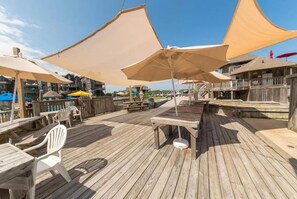 A wooden deck with outdoor seating, including tables and chairs, and large umbrellas providing shade. - BIS-Duck - 2BR/2BA - Great Pools and Beach! (Duck)
