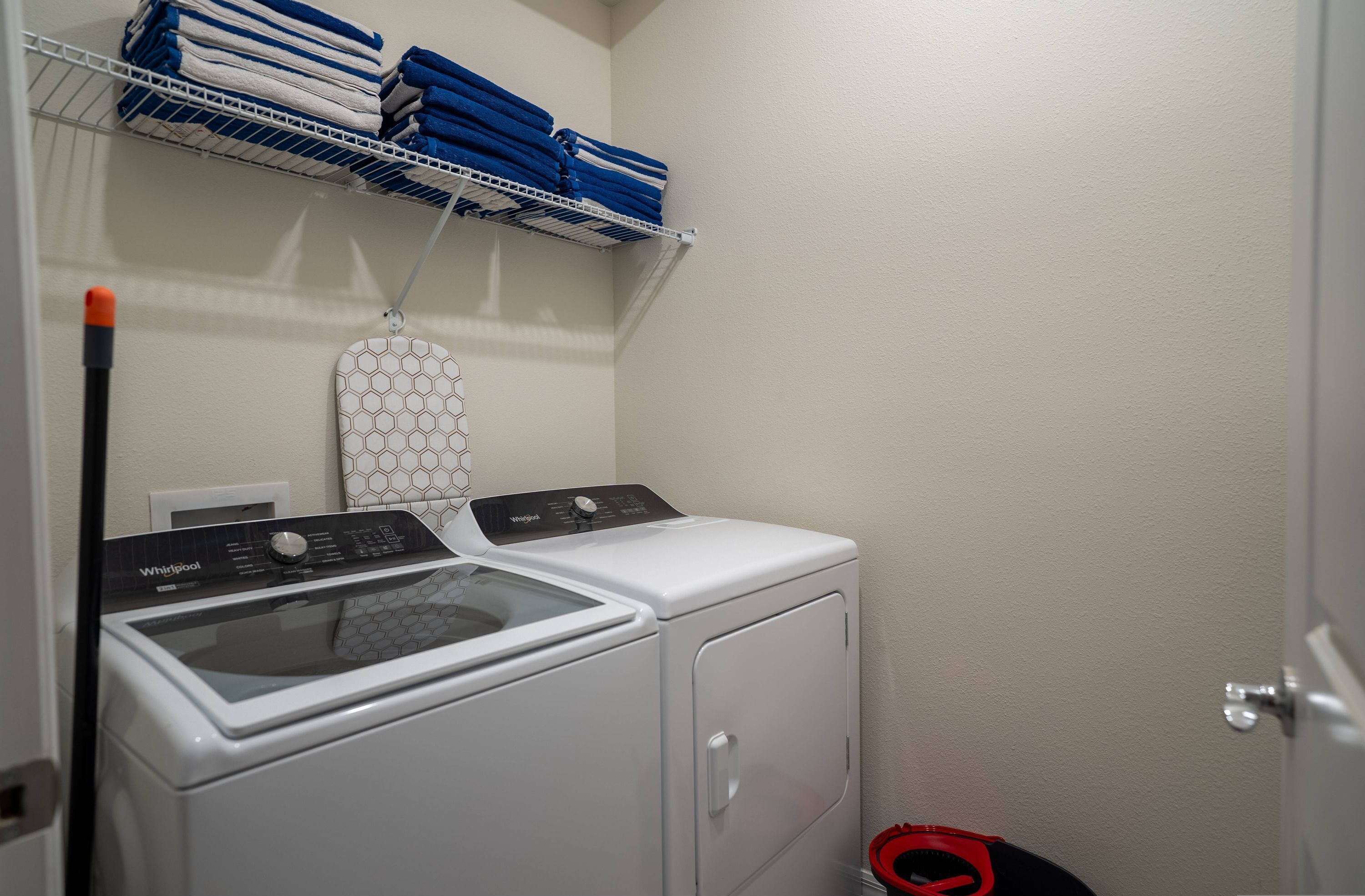 A laundry area with a front-loading washer and dryer, a metal shelf with folded towels, and a red and black cleaning tool on the floor.