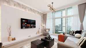 Ein modernes Wohnzimmer mit großem Wandfernseher, einer Couch, einem Couchtisch und Blick auf die Stadt durch große Fenster.