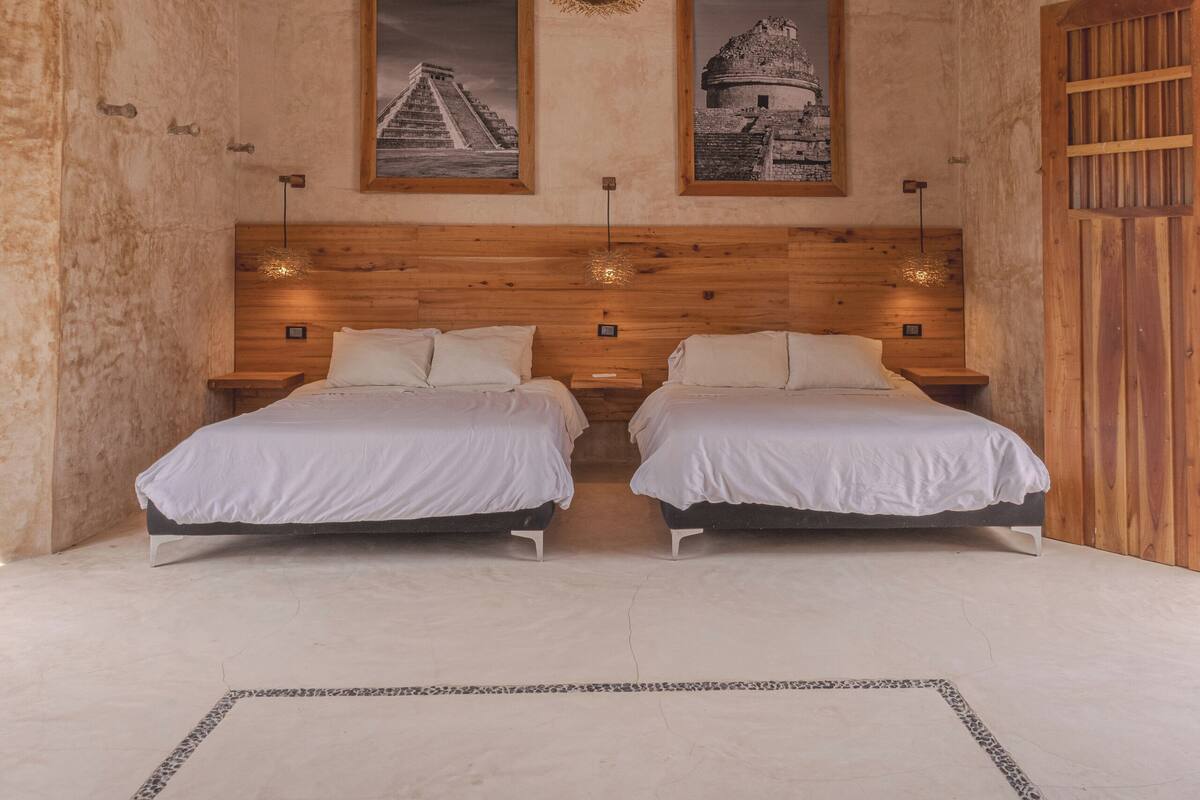 Een slaapkamer met een houten hoofdbord, twee bedden, een geweven plafondlamp en twee ingelijste foto’s aan de muur.