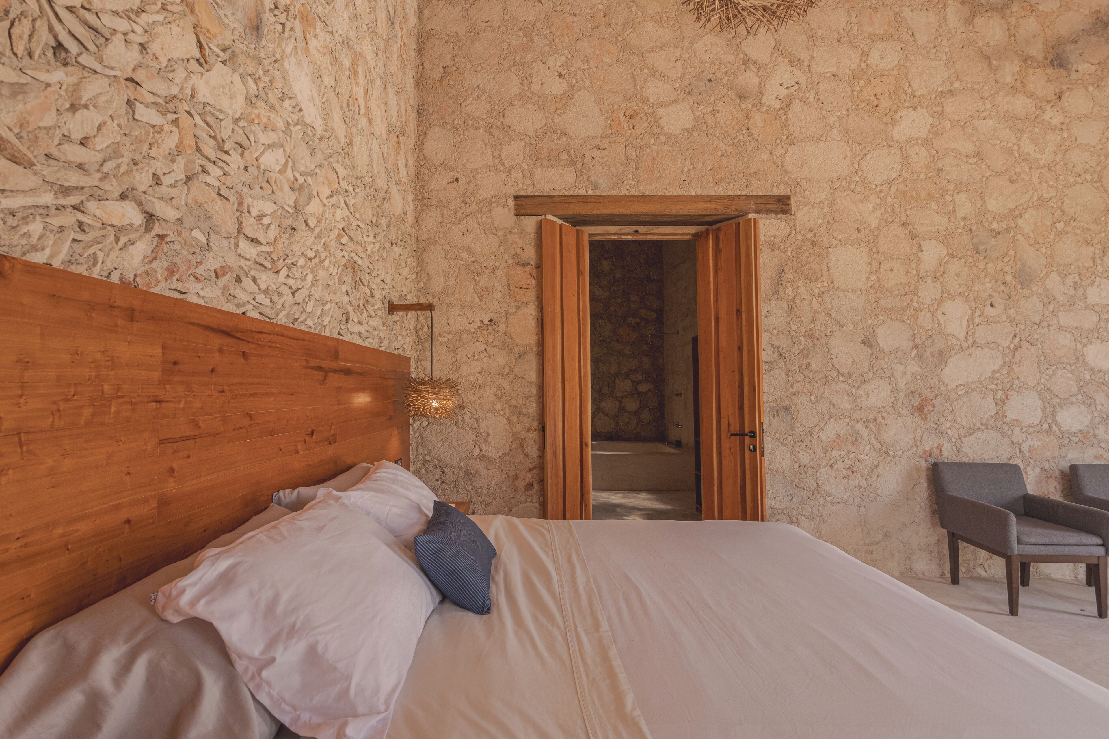 Un dormitorio con pared de piedra, cabecera de madera, cama con ropa blanca, mesita de noche, lámpara de pared y puerta que da al baño.