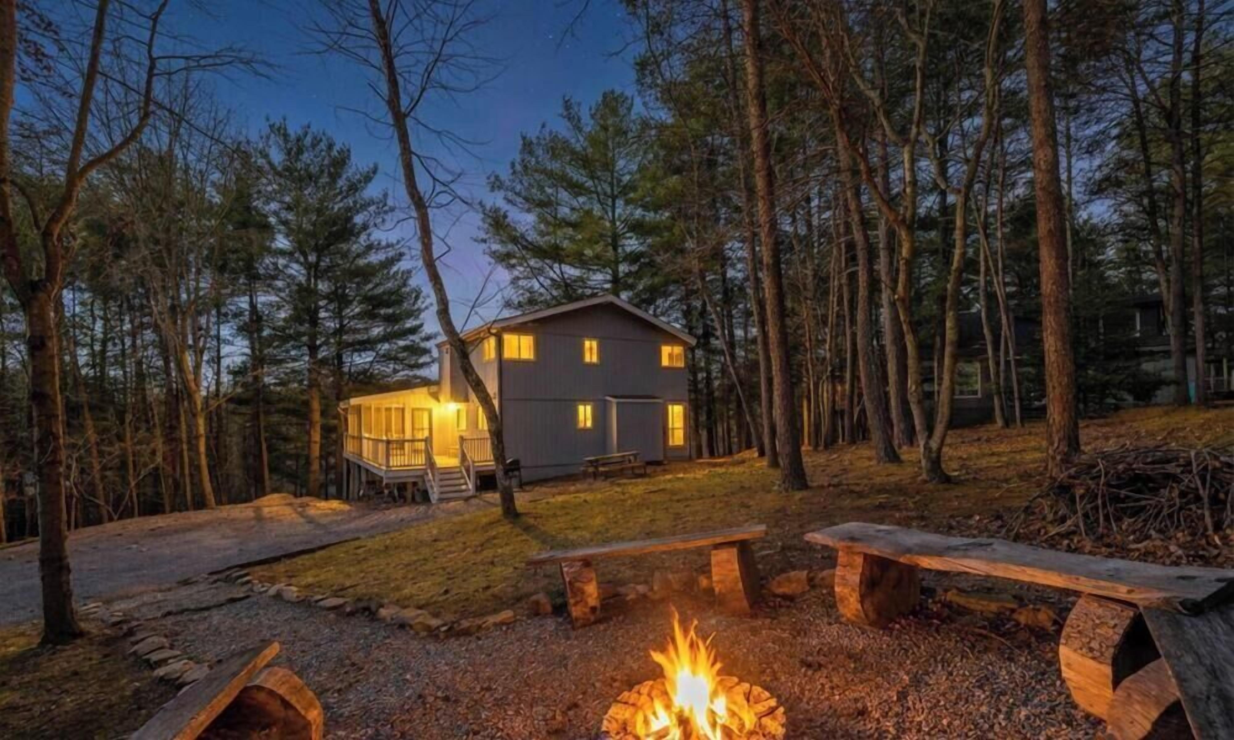 A cabin with a fire pit, surrounded by trees, and a bench in the foreground.
