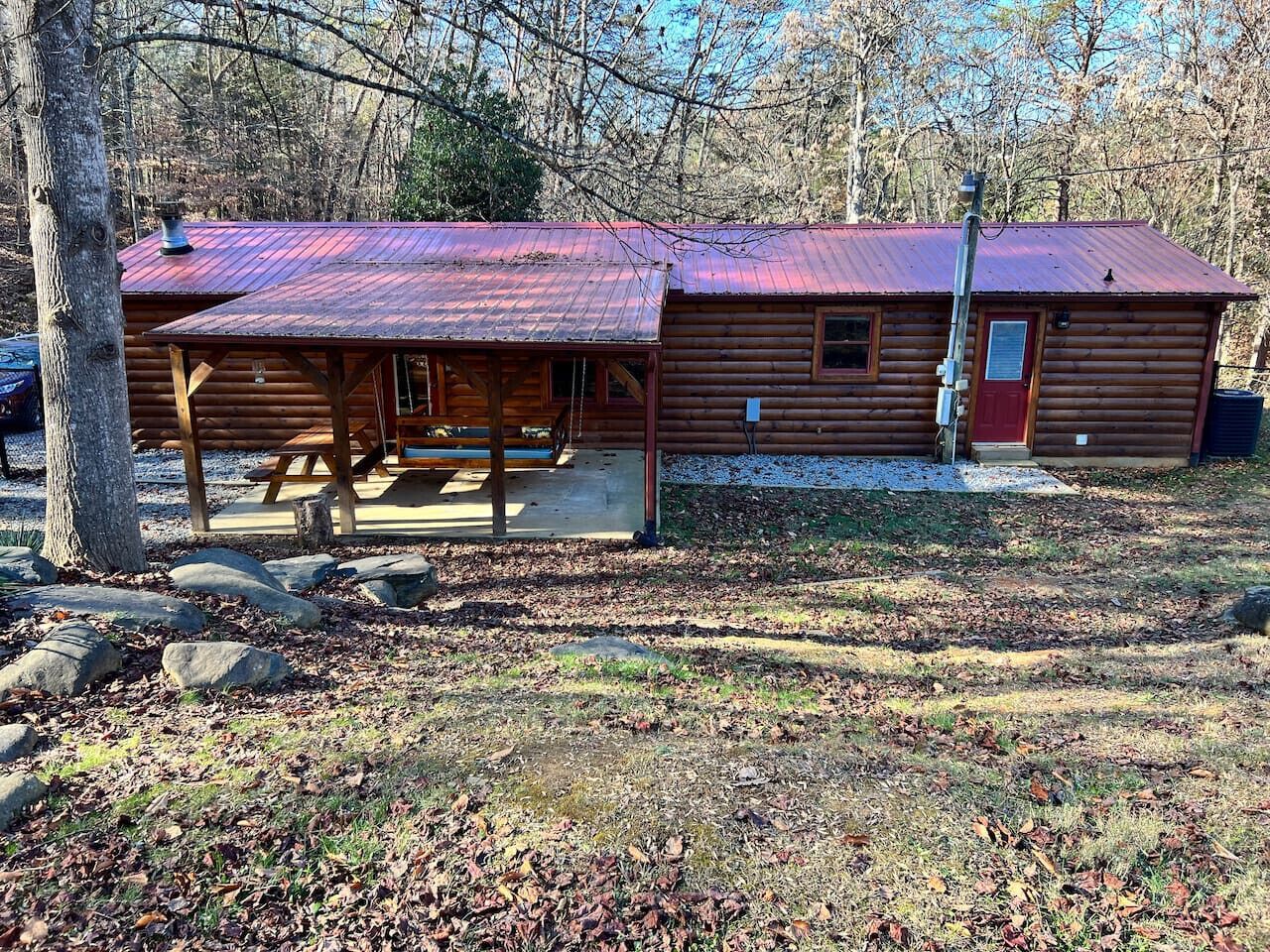 Una baita rustica con un portico coperto e un tetto rosso, circondata da alberi e un vialetto sterrato.