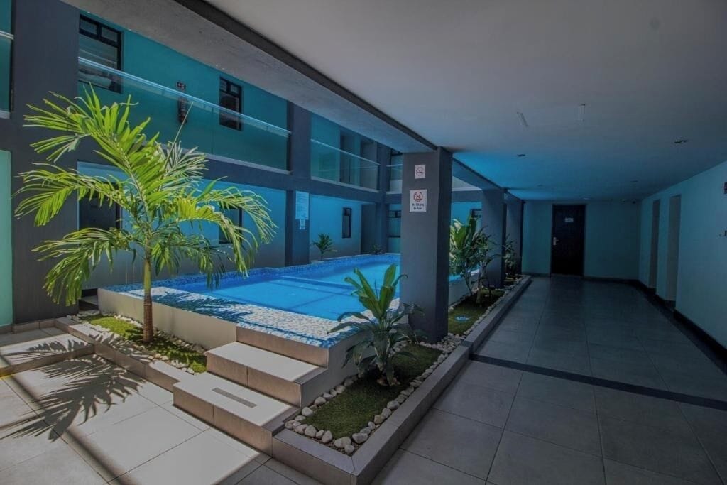 A modern indoor swimming pool with a glass enclosure, surrounded by a tiled walkway and small palm trees.