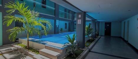 A modern indoor swimming pool with a glass enclosure, surrounded by a tiled walkway and small palm trees.