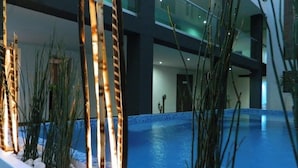 Una piscina interior amb vora de rajola, envoltada d'altes plantes i un edifici modern al fons.