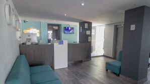 Un área de recepción con un mostrador, una computadora, un televisor y una zona de estar con sillas color turquesa.