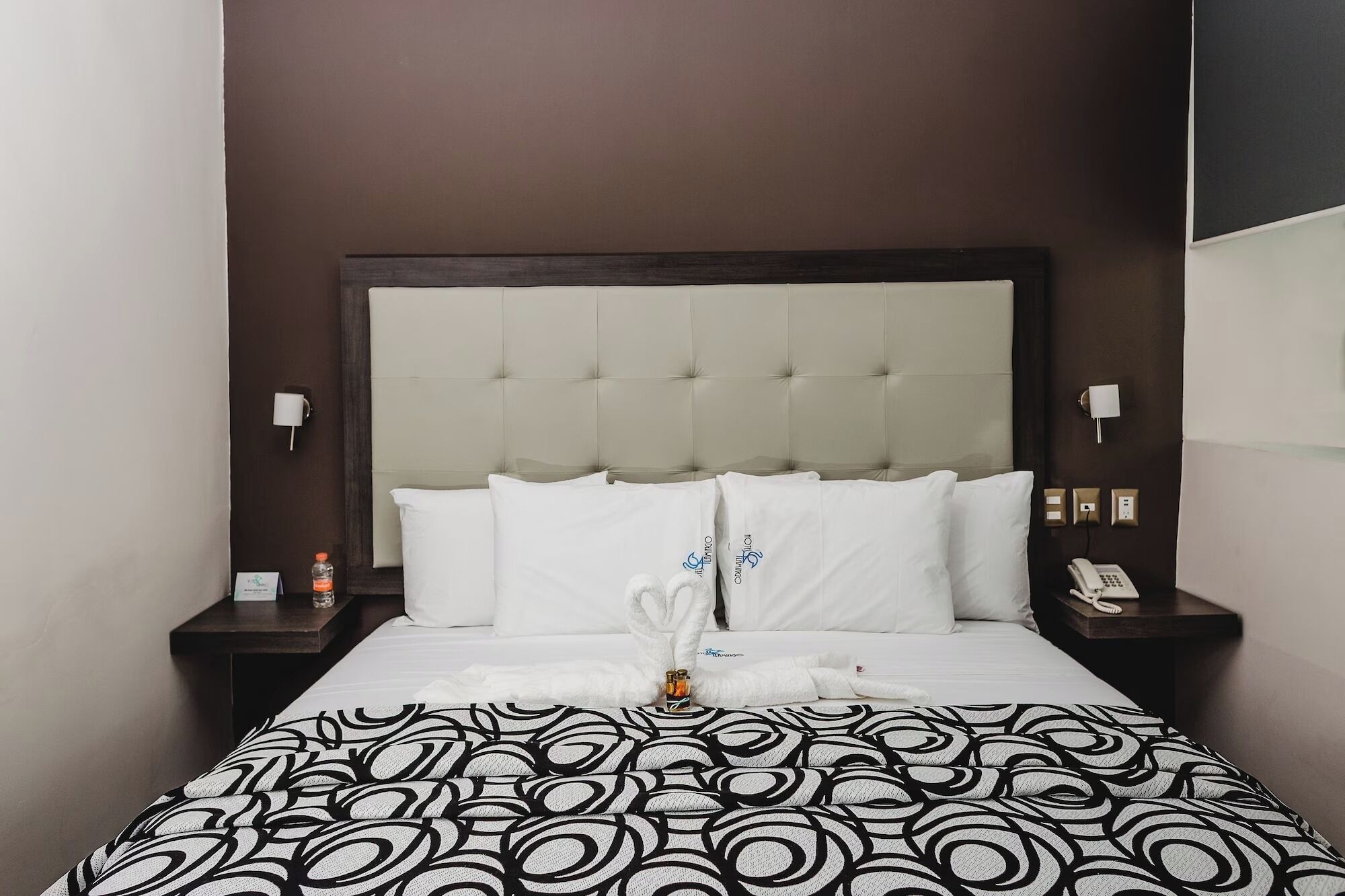 Een hotelkamer met een gestoffeerd hoofdeinde, witte beddengoed, een geborduurd plaid, een fles water en een nachtkastje telefoon.