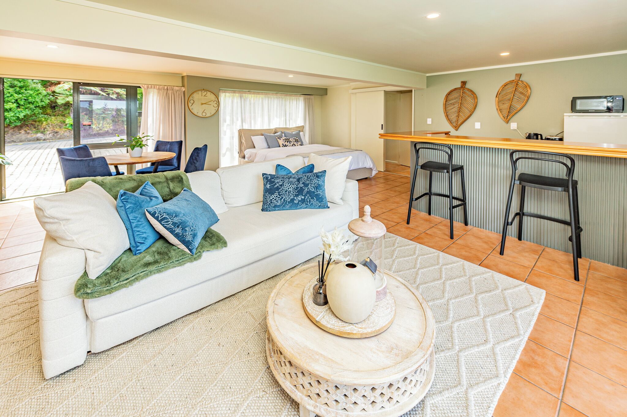 A modern living room with a white sofa, blue cushions, a round coffee table with a vase, and a kitchen area with a bar and two stools.