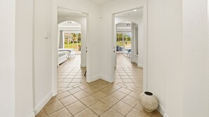 A hallway with white walls and tiled flooring leading to a living area with a sofa and a view of a garden. - Beautiful Townhouse with Golf Course Views, Near Attractions (Kissimmee)
