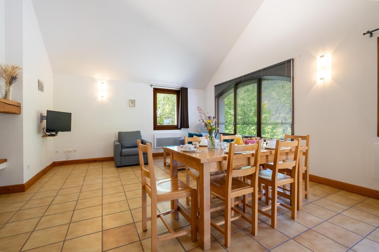 Un coin repas avec une table et des chaises en bois, un canapé gris et un téléviseur fixé au mur.