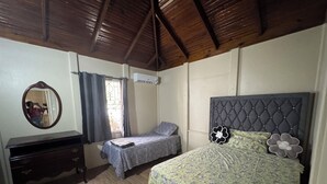 Een slaapkamer met een houten plafond, een bed met een bloemenbedekking, een nachtkastje, een dressoir, een spiegel en een raam met gordijnen.