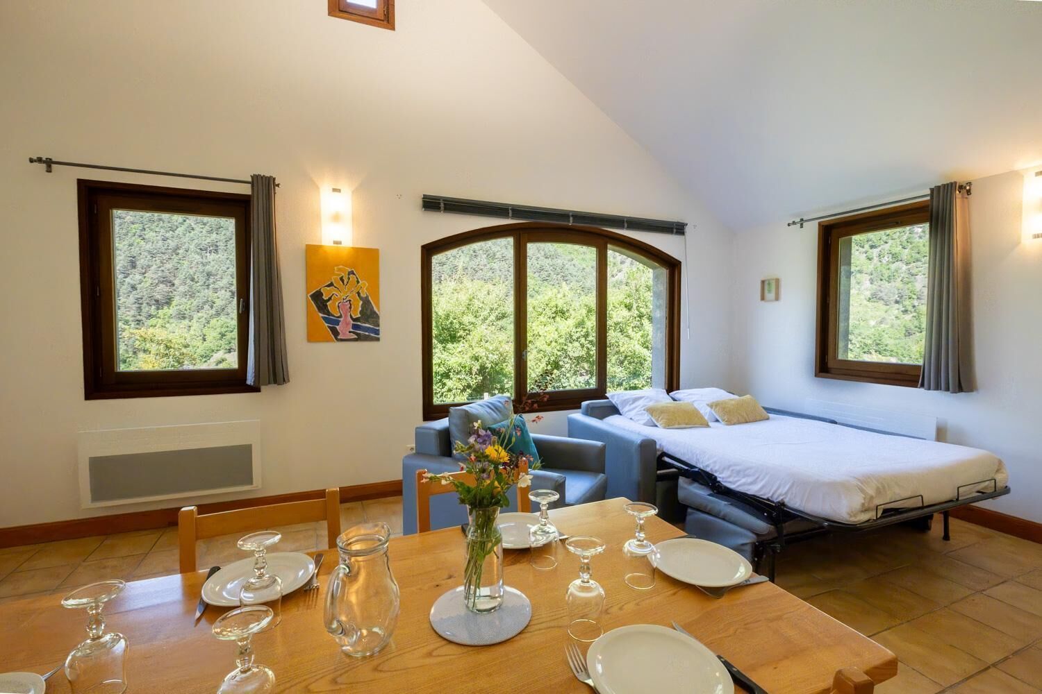 Un dormitorio con cama, sofá, mesa de comedor para dos personas y ventana con vista a árboles.