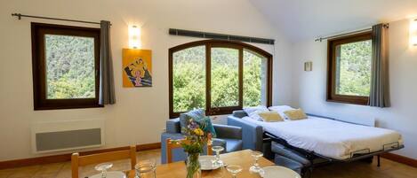 Un dormitorio con cama, sofá, mesa de comedor para dos personas y ventana con vista a árboles.
