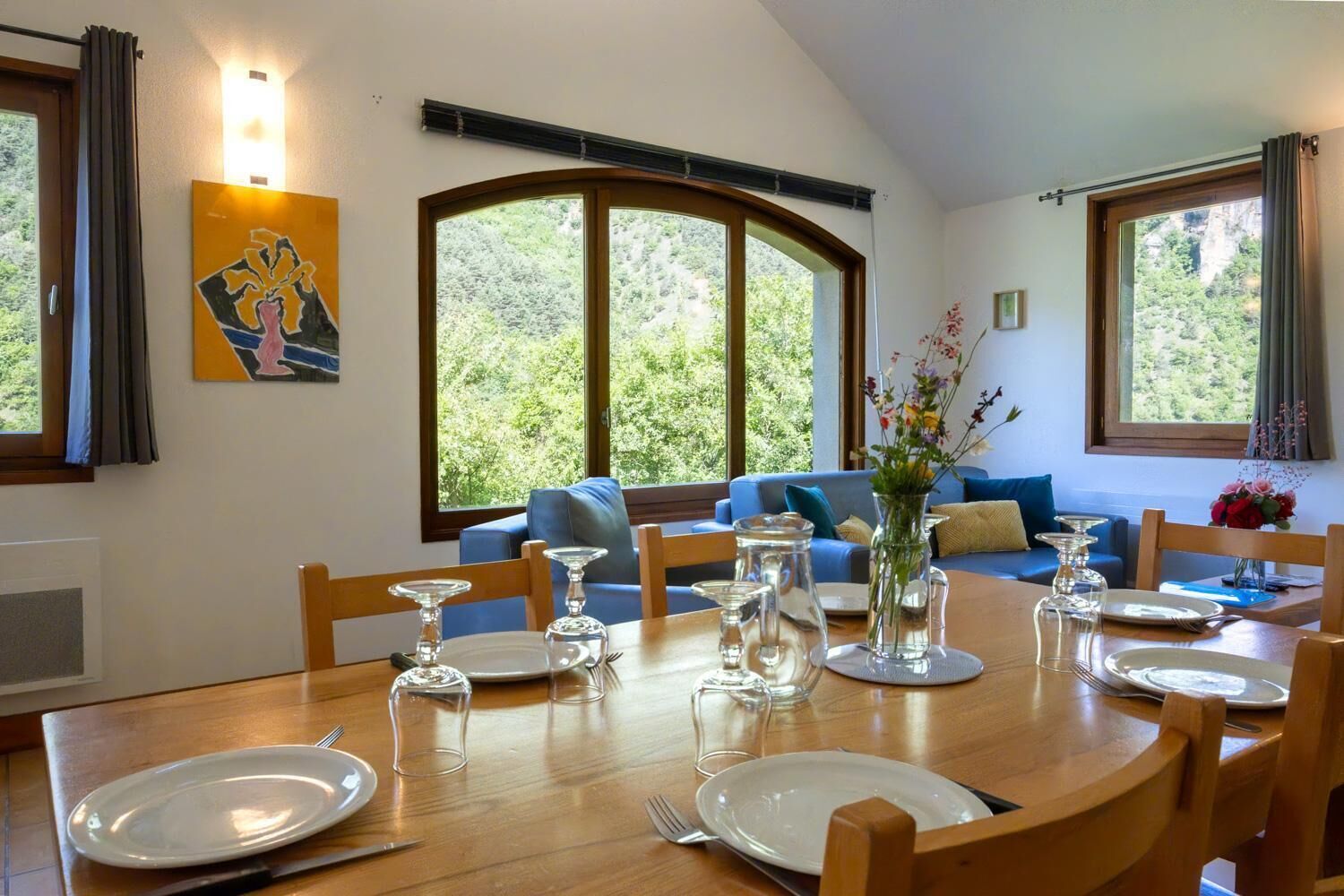 Comedor con una mesa de madera dispuesta para cuatro personas, rodeado de sofás azules y un ventanal con vistas a zonas verdes.
