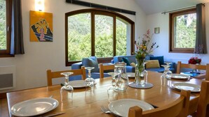 A dining area with a wooden table set for four, surrounded by blue sofas and a large window with a view of greenery. - Gîte 4/6 pers. Premium Plus at Castel de la Peyre (LES VIGNES)