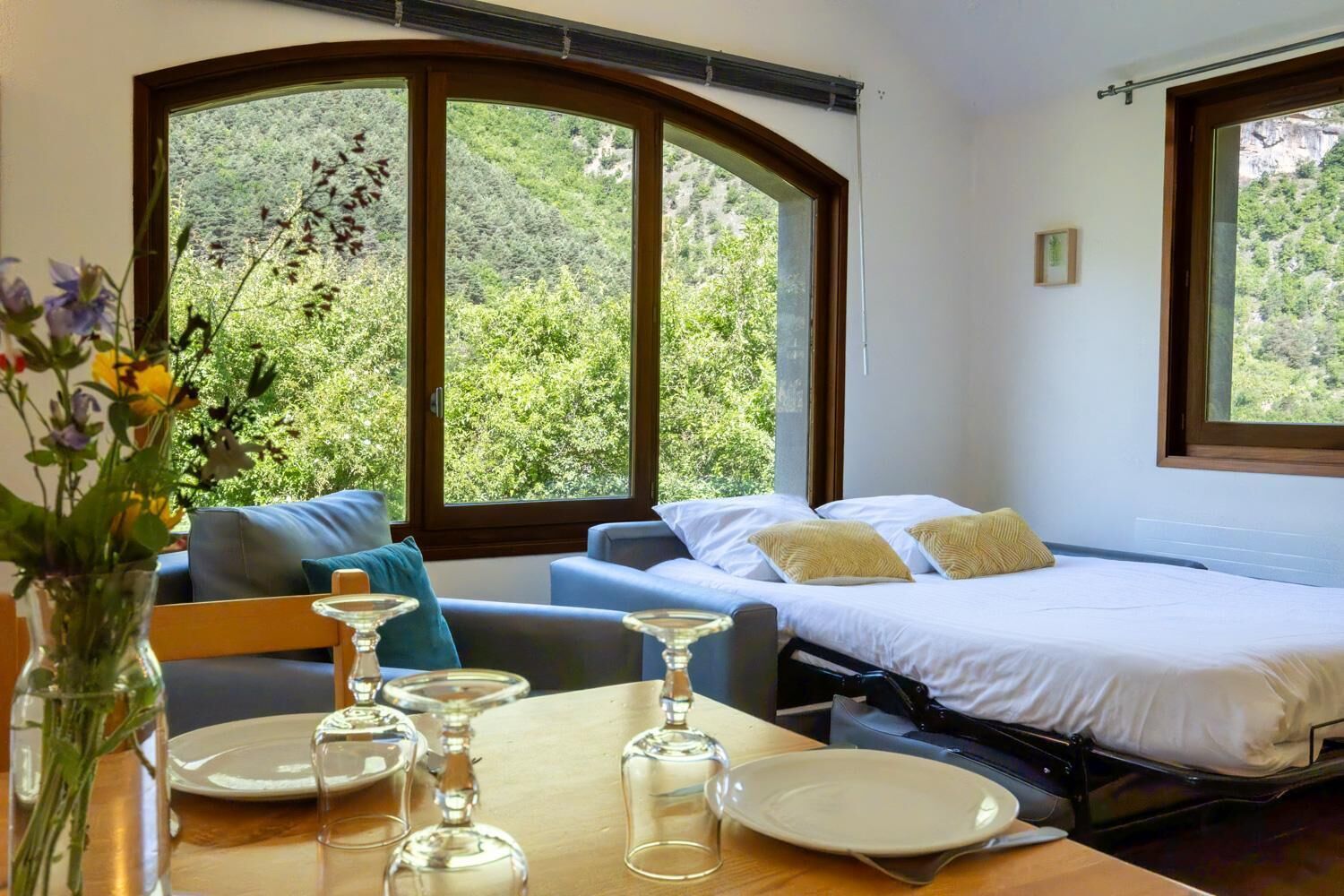 Un dormitorio con cama, sofá, mesa de comedor con platos y vasos, y ventanas amplias con vistas a la vegetación.