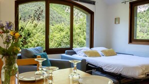 A bedroom with a bed, a sofa, a dining table with plates and glasses, and large windows with a view of greenery. - Gîte 4/6 pers. Premium Plus at Castel de la Peyre (LES VIGNES)