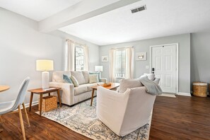 Un salon avec un canapé, un fauteuil et une table basse. On aperçoit une lampe, un tapis et une porte au fond.