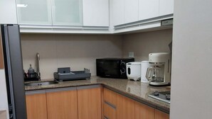 Et køkken med hvide skabe, en sort mikrobølgeovn, kaffemaskine og køleskab.