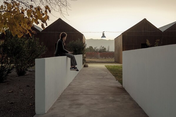 A person sitting on a white wall in a modern outdoor area with a concrete pathway, surrounded by wooden structures and some greenery. - aguamiel (Maipu)