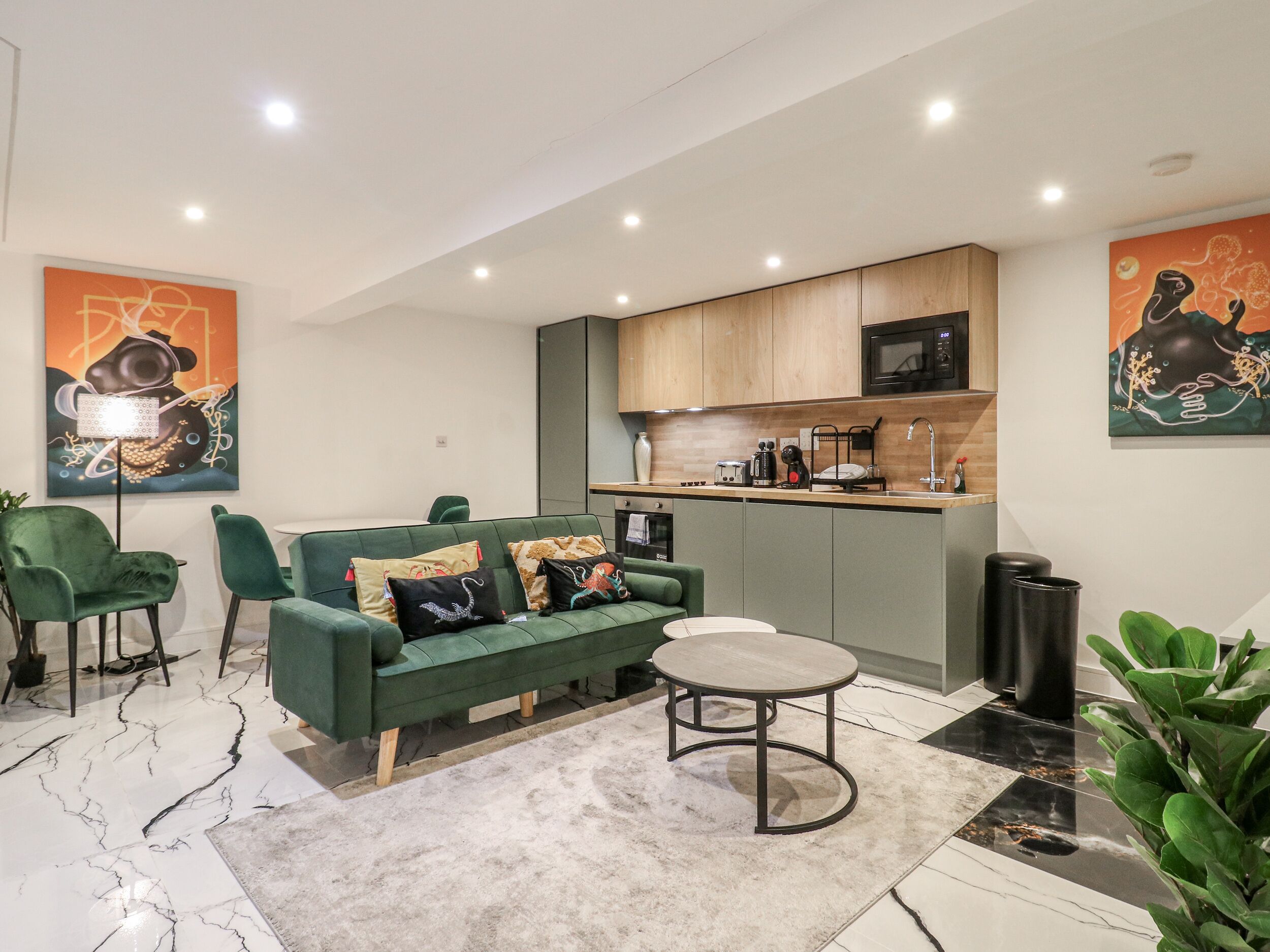 A modern living room with a green sofa, a round coffee table, and a kitchen area with wooden cabinets and a microwave.