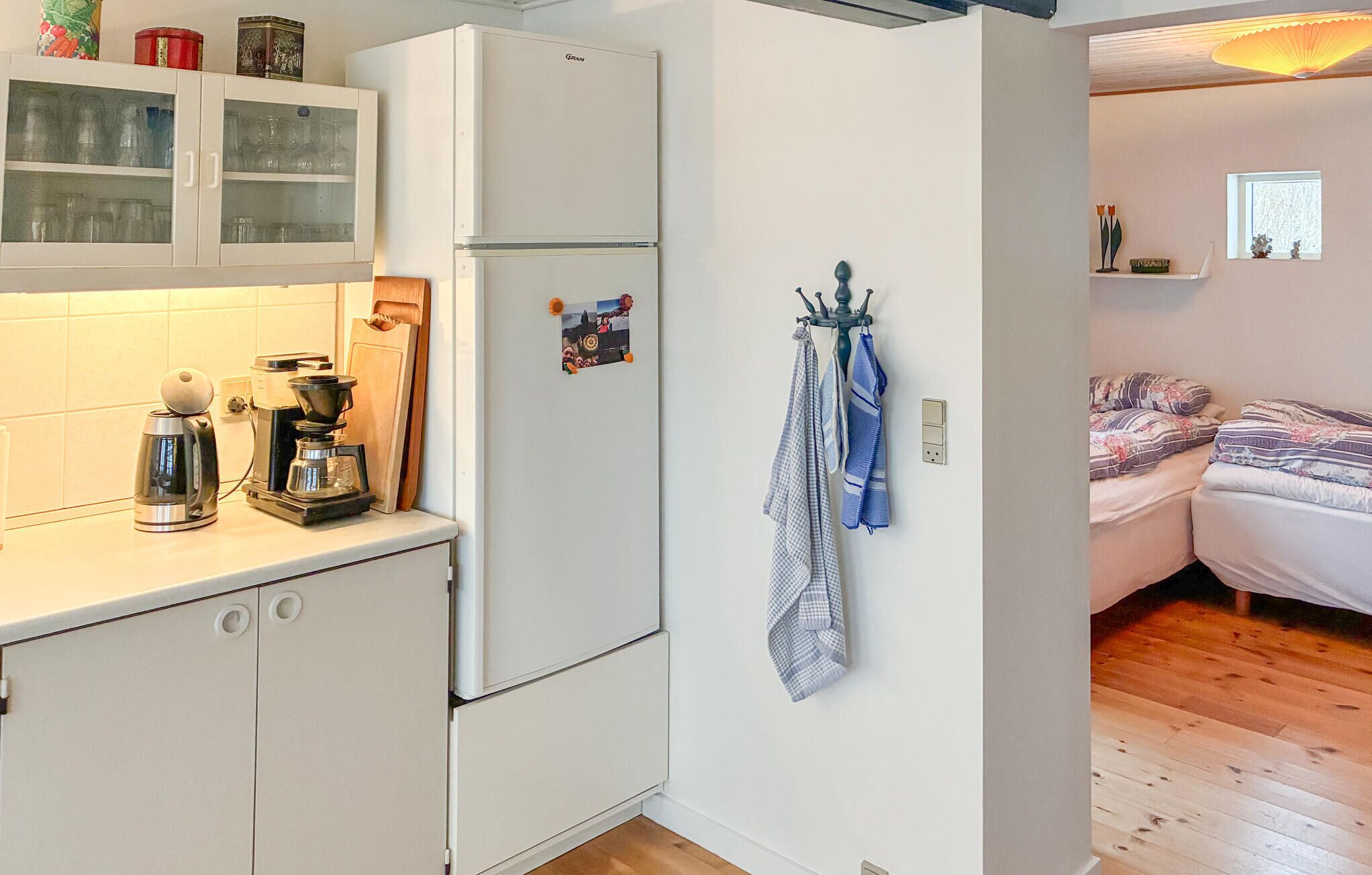 Cuina compacta amb nevera, cafetera i armaris. Es veu un llit amb llençols estampats a l'habitació propera.