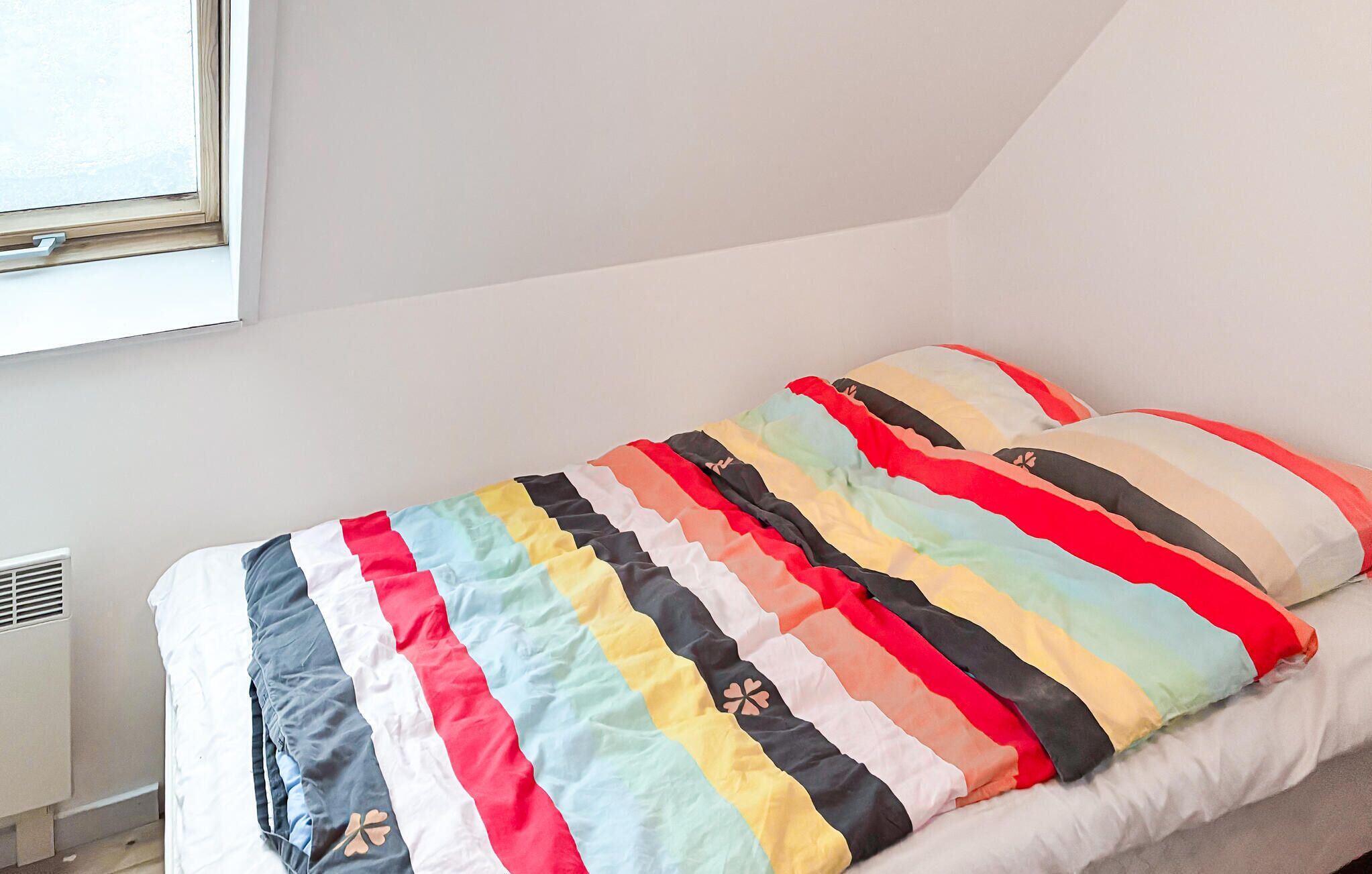 A single bed with a colorful striped blanket and matching pillows in a room with white walls and a window.