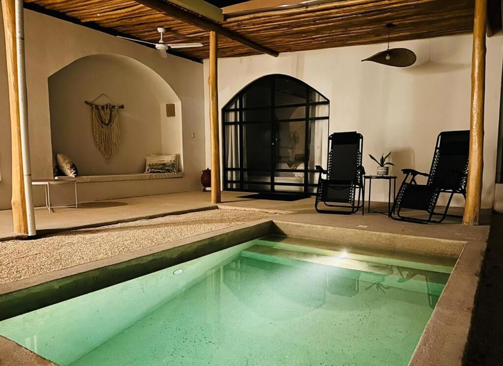 Una piscina interna con soffitto in legno, un’area relax con una panca e cuscini, e due poltrone nere.