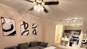 Ein Wohnzimmer mit einem Ecksofa, einem gemusterten Teppich, abstrakter Wandkunst, einem Deckenventilator und einer Küche im Hintergrund.