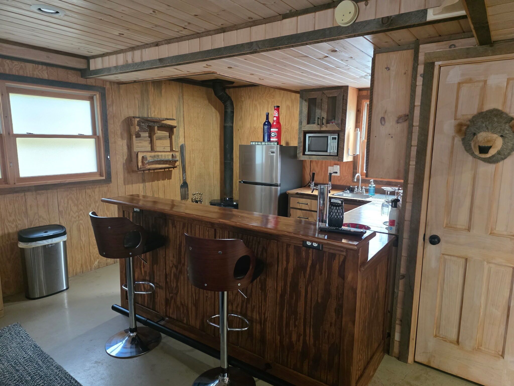 A kitchen with wooden paneling, a stainless steel refrigerator, a microwave, and a bar area with stools.