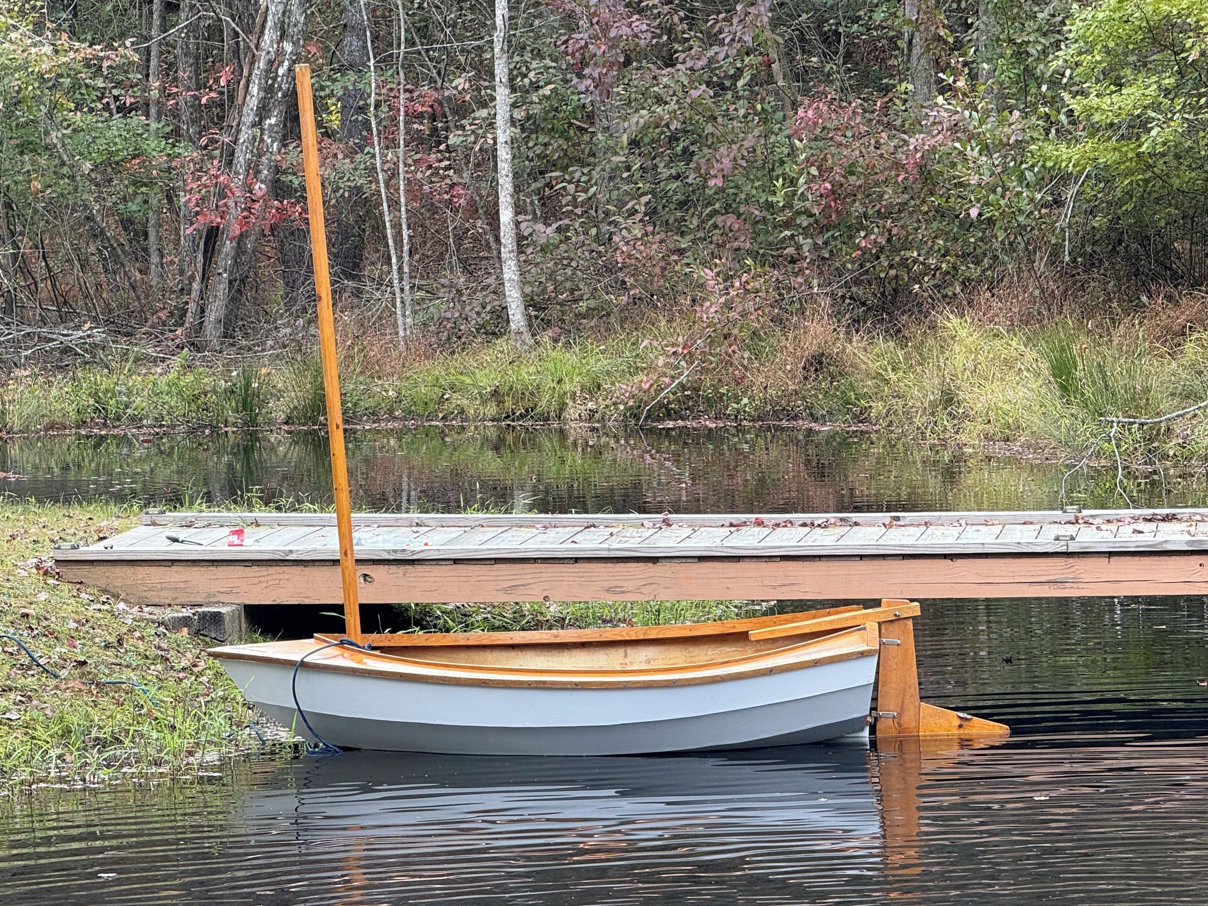 A small boat docked at a wooden pier extending into a calm lake, surrounded by dense trees and greenery.